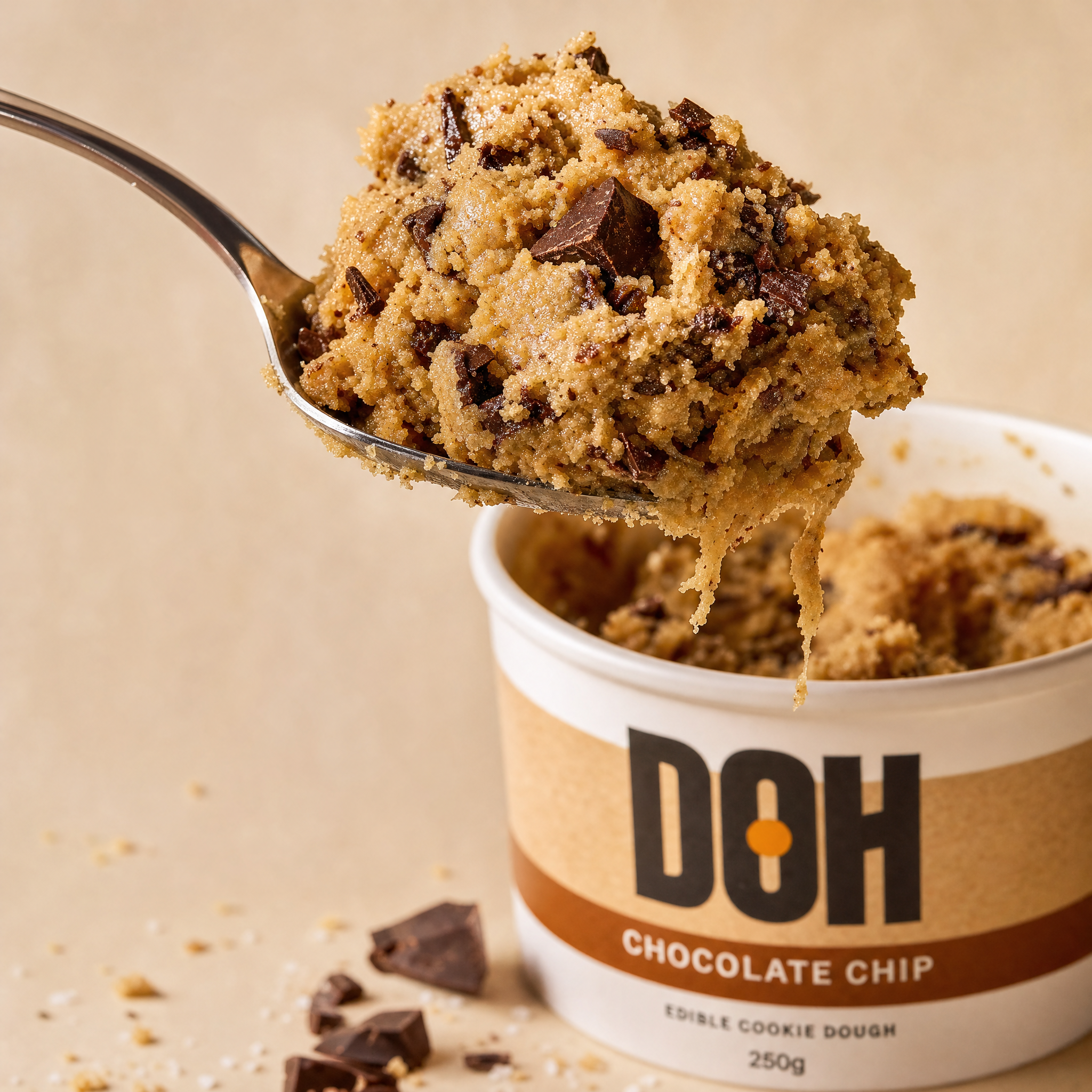 Close-up macro: spoon lifting chocolate chip cookie dough out of open DOH tub