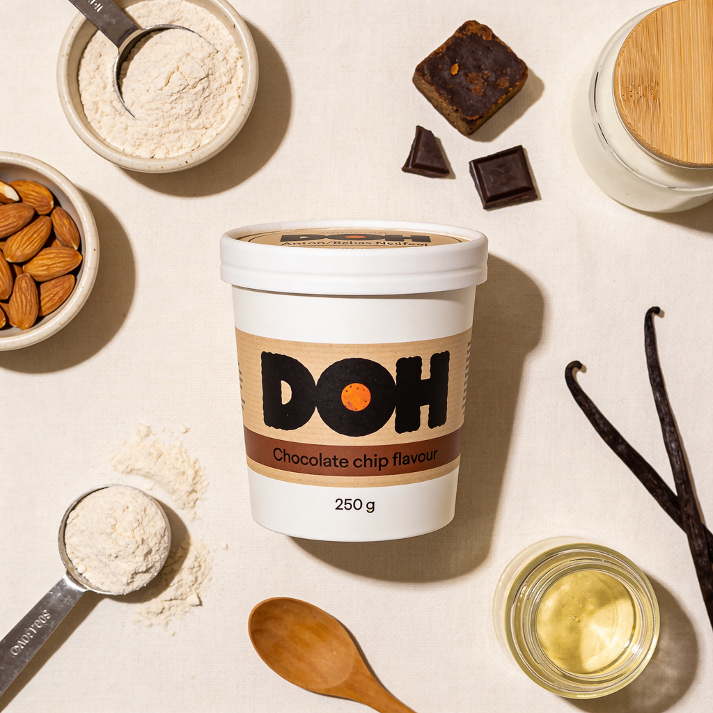 Overhead ingredient flatlay: DOH tub with almond flour, whey protein, jaggery, dark chocolate